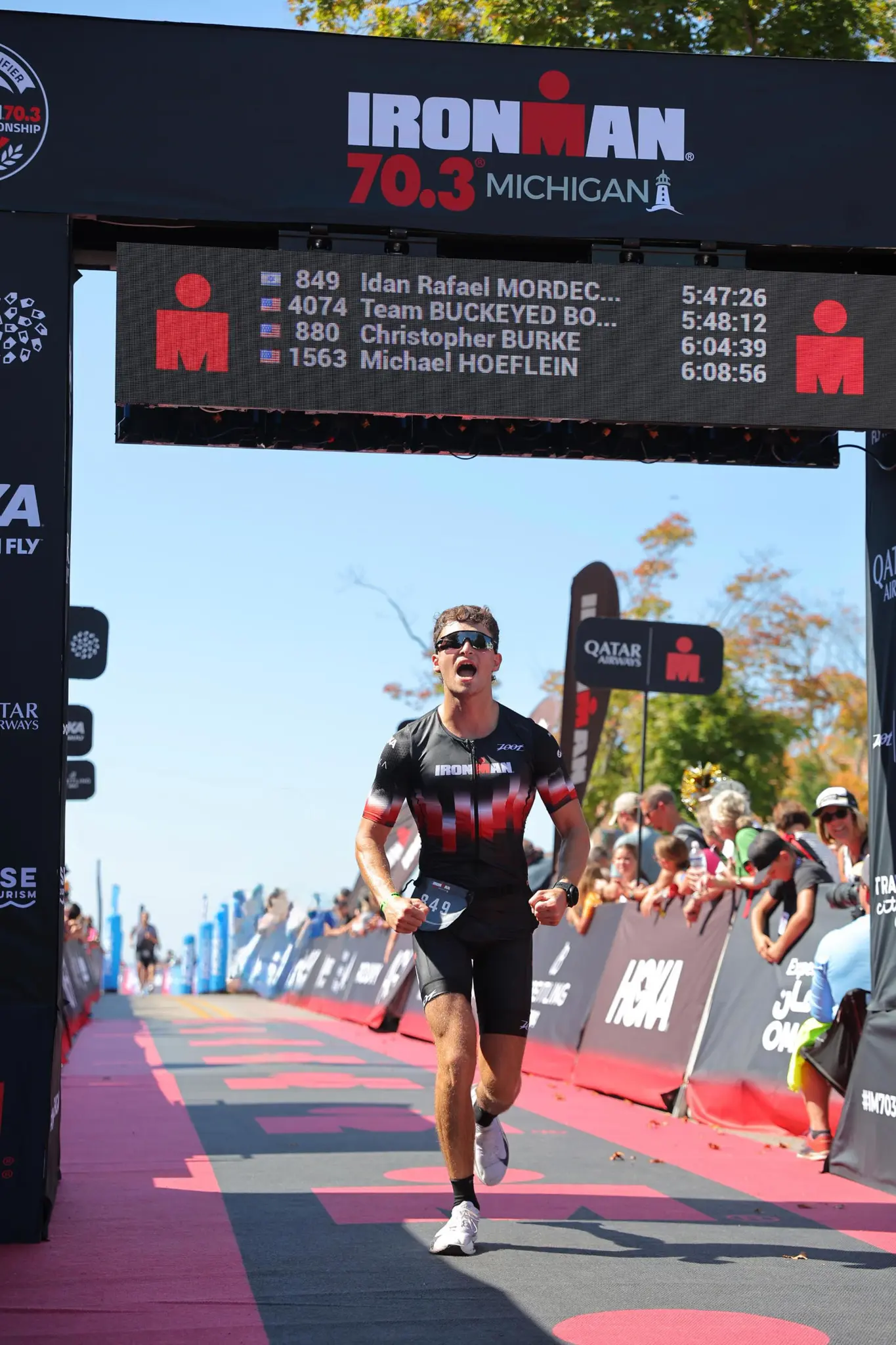 עידן רפאל מרדכי חוצה את קו הסיום של תחרות IRONMAN 70.3 מישיגן, לוח התוצאות מעליו מציג את שמו בזמן 5:47:26